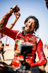 泥だらけのオフロードライダーが、青空の下で両手を上げて満面の笑みを浮かべ、勝利や達成感を喜ぶ