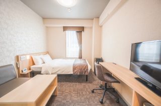 白い寝具のベッド、デスクとテレビ、ソファが配置された清潔感のあるホテルの客室です。