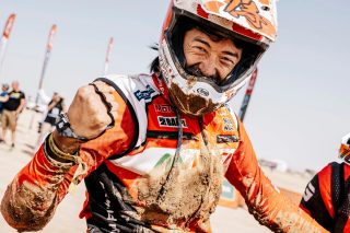 泥だらけのレーシングスーツとヘルメットを着用した男性が、満面の笑みでガッツポーズ