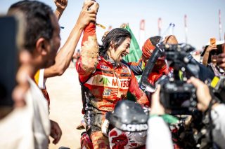 泥だらけのレーシングスーツを着たレーサーが、周囲の人々と共に勝利を喜び、満面の笑みで腕を上げる