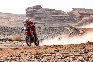 砂漠のような荒涼とした地形を、赤いレーシングスーツを着たライダーがホンダのオフロードバイクで疾走