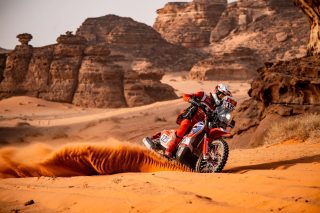 赤いオフロードバイクに乗ったライダーが砂漠を疾走し、後方には砂煙が舞い上がる