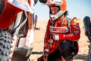 赤いレーシングスーツとヘルメットを着用した日本人ラリーライダーが、砂漠の景色の中でオフロードバイクの傍らにしゃがみ込む