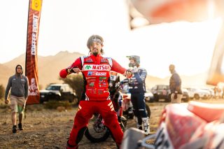 夕暮れ時の砂漠地帯で、赤いレーシングスーツを着た日本人ライダーがポーズ