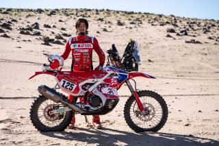 砂漠の広がる場所でオフロードバイクの横に立つ赤いレーシングスーツのライダー