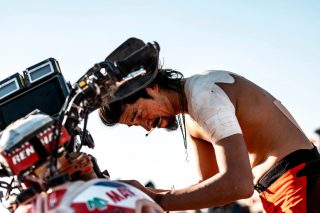 肩に包帯を巻いたライダーが、激しいレース中にバイクに寄りかかり、疲労困憊の様子でうつむいている