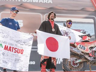 ダカールラリーの表彰台で、日本人選手が日の丸の旗と応援メッセージが書かれたバナーを掲げ、満面の笑みを浮かべる