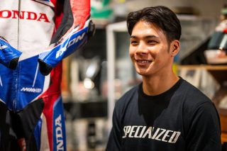 笑顔を見せる若い男性が、赤、白、青のホンダMotoGPレーシングスーツの隣に立っています。男性はSPECIALIZEDのTシャツを着用しており、屋内でのリラックスした雰囲気が伝わります。