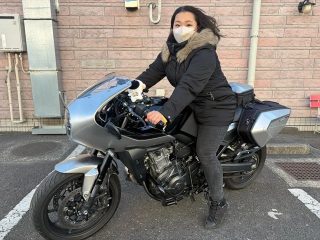 マスクと冬用ジャケットを着用した女性が、銀色のバイクにまたがっている