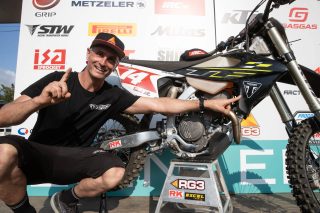 モトクロスイベントで、笑顔の男性がトライアンフのオフロードバイクを指差している。背景には多くのスポンサーロゴがあり、男性はライダーのステファン・グランクイスト。