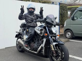 ヘルメットとライダースーツを着用した男性が、黒い大型バイクに乗り、両手でVサイン