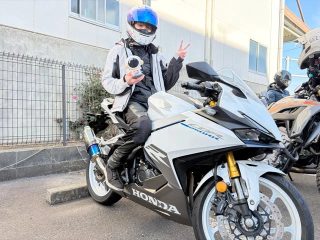 ヘルメットとライディングウェアを着用した人物が、白いホンダCBR250RRに跨り、宇宙飛行士のフィギュアを持ちながらピースサインをしている