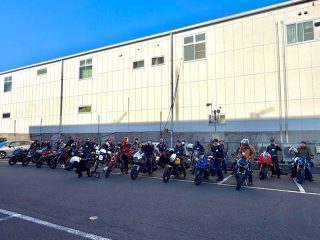 多くのライダーたちが様々なオートバイと共に、建物の前で集合写真を撮っている