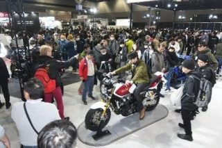 多くの人々で賑わう屋内イベント会場で、展示された赤と白のオートバイに男性が跨っている