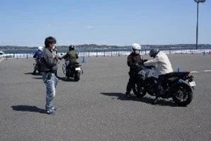 青空の下、アスファルトの広場で数人がバイクの運転練習をしている様子