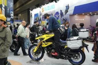屋内の展示会場で、一人の男性が黄色のスズキ製アドベンチャーバイクに跨っています