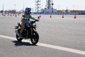 ヘルメットを着用した人が、オレンジ色のパイロンが並べられたアスファルトの広場でバイクに乗って走行している様子