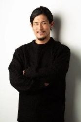 黒いセーターを着て腕を組み、カメラを見つめる髭の男性