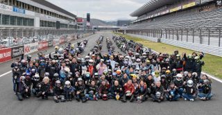 富士スピードウェイのコース上で、多数のバイクとヘルメットを着用したライダーたちが、楽しそうに集合写真を撮っています。背景には多くのバイクと観客席が見えます。