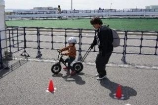 屋外の広場で、大人が後ろから補助棒を持って子供の自転車練習を手伝っている様子