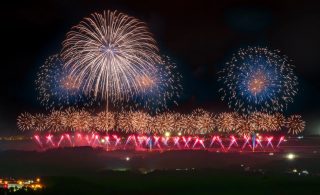 夜空を彩る壮大な花火大会の様子。金色や青色の大きな打ち上げ花火が輝き、地上近くには赤と青の光の演出も加わり、幻想的で華やかな祝祭の雰囲気を醸し出しています。