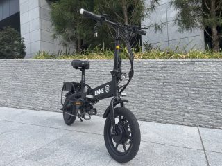 黒い折りたたみ式電動アシスト自転車が、モダンな建物の前にある石畳の歩道に停まっている様子