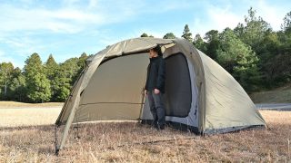 広々としたテントまたはタープの中に立つ男性の姿を捉えた一枚。晴れた日の屋外で、キャンプやアウトドアレジャーを楽しむ様子が伺えます。