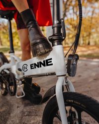 白い折りたたみ電動自転車に乗る人物の足元を捉えた画像です。秋の風景の中で、黒いブーツを履いた足がペダルに置かれています。自転車のフレームには「ENNE」というロゴが見えます。