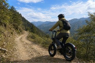 ヘルメットとリュックを背負った男性が、緑豊かな山々が広がるオフロードの道を自転車で走行している様子