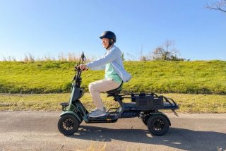 ヘルメットを着用した男性が、晴れた日に緑の丘と青空を背景に、カーキ色の三輪電動スクーターに乗って道を走行している様子