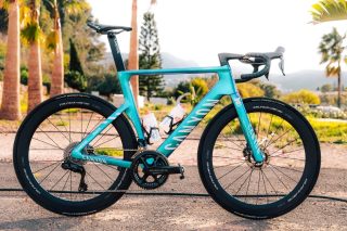 ターコイズブルーのCANYONロードバイクが屋外に駐輪されている画像です。DURA-ACEホイールやSHIMANOコンポーネントを装備した高性能なモデルで、背景にはヤシの木と山が見え、穏やかな気候の場所でのサイクリングシーンを想起させます。
