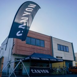 CANYONのロゴが入った大きな旗とテントが設置された屋外イベント会場の様子。テントの下には自転車が並び、スタッフらしき人々が活動している。建物には「R-ing.TCR」の表示もある。