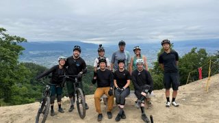 山頂でマウンテンバイクを楽しむ9人のグループが記念撮影をしている。
