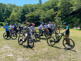 晴れた日、森を背景にした芝生の上で、ヘルメットを着用した人々がマウンテンバイクと共に集まっています。