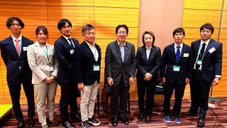 ビジネススーツを着用した日本人男女9名が、会議室のような場所で笑顔で並んで写る集合写真