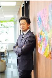 現代的なオフィス空間で、ダークスーツを着た日本人ビジネスマンが腕を組み、壁に寄りかかっています。隣にはカラフルな抽象画が飾られ、男性は自信に満ちた表情でカメラを見ています。