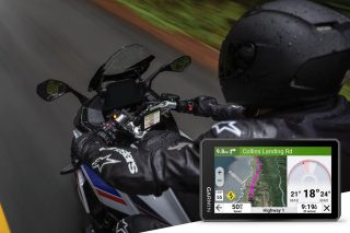 雨の中、バイクに乗るライダーがGarminのナビゲーションシステムを使用している様子。画面には目的地へのルート、速度制限、気温などが表示され、海岸沿いの道路を走行中。ライダーはAlpinestarsのギアを着用しています。