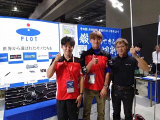 展示会ブースで3人の男性が笑顔でサムズアップしています。