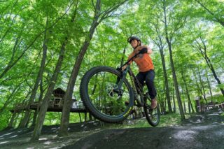 新緑がまぶしい森の中で、マウンテンバイクに乗った人物が笑顔でジャンプしている瞬間