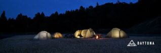 夜の広々とした野原で、いくつかのテントが明かりを灯し、焚き火が燃えています。奥には星が輝く夜空と暗い森が広がり、手前にはバイクも見える、静かで美しいキャンプの風景です。