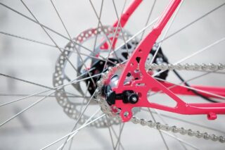 鮮やかなピンク色の自転車の後輪部分をクローズアップした画像です。銀色のディスクブレーキ、細いスポーク、駆動チェーンが詳細に写っており、自転車の機械的な美しさを捉えています。