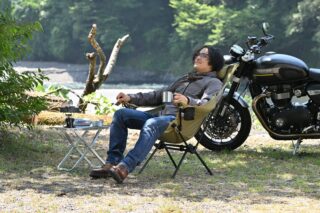 自然の中で、男性がバイクの横でキャンプチェアに座り、リラックスしています。手にはマグカップを持ち、隣のテーブルにはポータブルストーブが置かれ、川を眺めながら休憩しているようです。
