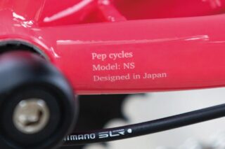 赤い自転車のフレーム部分をクローズアップした画像です。「Pep cycles Model: NS Designed in Japan」という文字と、SHIMANOのケーブルが写っています。日本でデザインされた自転車の細部を示しています。