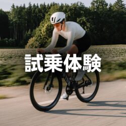 白いヘルメットとサングラスを着用した人がロードバイクに乗って舗装された道を走行しています。背景には木々や畑が広がり、画像には「試乗体験」という文字が重ねられています。