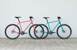 ピンクとターコイズブルーの2台の自転車が、白い壁を背景に並べられています。シンプルで清潔感のある構図が特徴です。