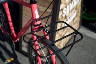 赤いフレームの自転車のフロント部分をクローズアップした写真です。黒いフロントラックが前輪の上に設置されており、タイヤのトレッドパターンも確認できます。