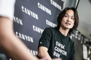 「CANYON」のロゴが並ぶ壁を背景に、黒いTシャツを着たアジア人男性が話している。Tシャツには「SWIM. CANYON. RUN.」の文字があり、スポーツ関連のイベントやインタビューの様子を捉えた一枚。