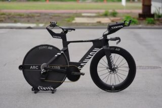 黒いCANYON製タイムトライアルバイクが舗装路に停車しています。空気抵抗を最小限に抑えた流線形のフレームデザインと、後輪のディスクホイール、前輪のディープリムホイールが特徴で、高性能な競技用自転車です。