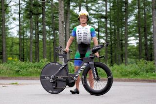 トライアスロンウェアとヘルメットを着用した男性アスリートが、黒いタイムトライアルバイクと共に森林を背景に立っている様子。競技前の準備やトレーニング風景を捉えた一枚。