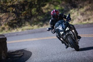 ヘルメットとライディングギアを身につけたライダーが、黒いスポーツバイクで舗装された道を深くバンクさせながらカーブを走行している